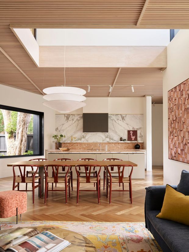 A refined open-plan dining and living space with a marble-backed kitchen, warm timber slatted ceiling with a large skylight or clerestory opening, herringbone timber flooring, and a long timber dining table surrounded by red-toned bentwood chairs. A dark sofa, mustard cushion, patterned rug, sculptural pendant, and large abstract artwork add colour and character.