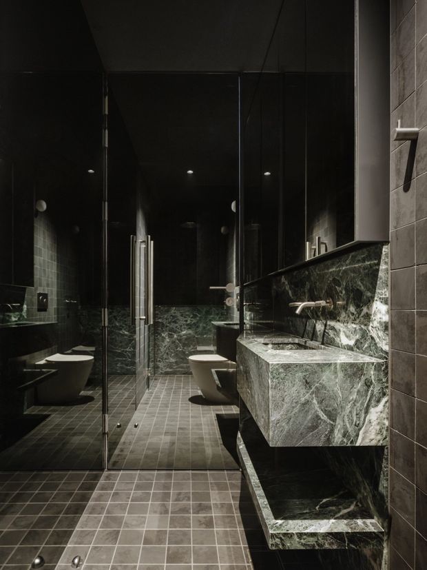 Dark, dramatic contemporary bathroom with a glass-enclosed shower, wall-mounted toilet, floating marble vanity, large mirror, and grey tiled surfaces. The space uses deep charcoal tones, richly veined green-grey stone, brushed metal fixtures, and recessed ceiling lighting.