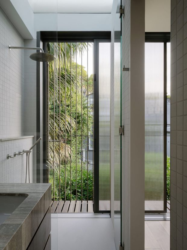 A contemporary bathroom with a walk-in shower area, white small-format wall tiles, dark grey stone vanity benchtop, and a frameless or minimally framed glass shower screen. Full-height glazed openings with dark aluminium frames and vertical security bars bring in greenery and daylight. A timber-look slatted shower floor contrasts with the pale tiled floor, while a rainfall shower and handheld fixture are visible.