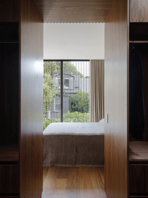 A compact bedroom viewed through a full-height timber wardrobe opening. The room features warm timber flooring, integrated dark and light wood cabinetry on both sides, a simple bed with textured neutral bedding, and a large window with vertical metal bars and beige curtains overlooking greenery and neighbouring buildings. The design is highly tailored and understated, with warm natural materials and clean contemporary detailing.