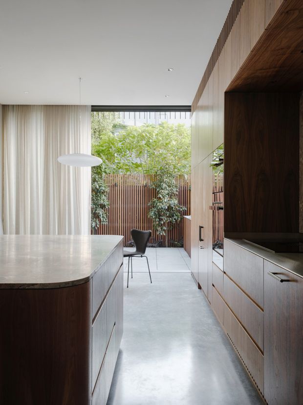 A refined contemporary kitchen with a large rounded-edge island, extensive warm timber cabinetry, pale stone benchtops, and a polished concrete floor. Full-height curtains line one side, while an open glazed area leads to a leafy courtyard with timber screening. Integrated appliances and a dark timber-lined niche add depth to the minimalist joinery.