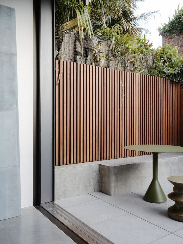 A contemporary courtyard or patio viewed from inside through a wide black-framed glazed opening. The outdoor space has large pale concrete or stone-look paving, a built-in grey concrete bench, a tall vertical timber privacy screen, dense tropical planting behind wire mesh, and a small olive-green round table with sculptural seating. A brick wall is partially visible at the upper right.