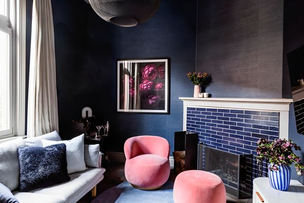 A richly styled living room with deep navy walls, a white-painted fireplace mantel, and a glossy navy brick-tile fireplace surround. A pale sofa with dark and light cushions sits beside two rounded blush-pink upholstered chairs, while a framed floral artwork, curtains, flowers, and a sculptural pendant add decorative detail. The room is brightened by a large window at left and has a refined, dramatic atmosphere.