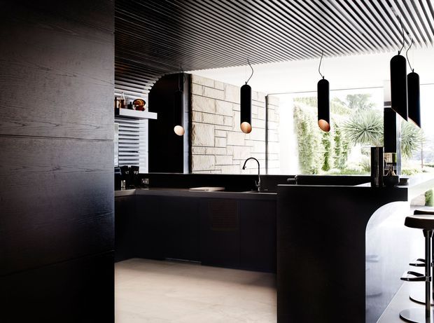 Dark, sophisticated contemporary kitchen with black cabinetry and island, a pale stone feature wall, expansive glazing, and a striking linear slatted ceiling. Black pendant lights hang above the island, while timber stools and greenery are visible beyond the kitchen.