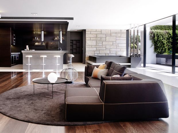A bright, open-plan contemporary living room with a large dark sectional sofa arranged around a low round coffee table and rug. The space includes a sleek kitchen with a stone island and bar stools, dark timber cabinetry and ceiling lining, a large rectangular stone feature wall with an integrated fireplace, polished timber flooring, floor-to-ceiling glazing, and a vertical green wall visible near the terrace.