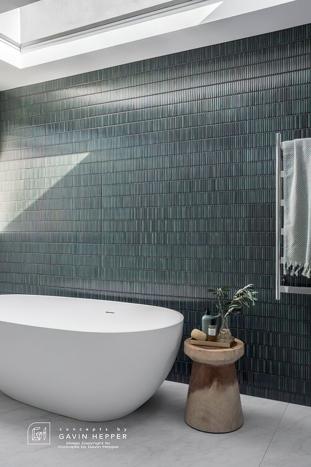 Contemporary bathroom featuring a freestanding white oval bathtub against a full-height dark vertically ribbed tile feature wall. A skylight brings strong natural light into the space, with a timber pedestal side table holding toiletries and greenery. A textured wall hanging is partially visible at right, and the floor is a pale grey finish.