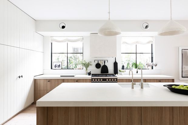 Bright, spacious contemporary kitchen with a large white island, warm timber cabinetry, white walls and benchtops, black-framed windows, pendant lights, and a freestanding range beneath a simple white rangehood. The clean, light-filled design has subtle coastal and Scandinavian influences.