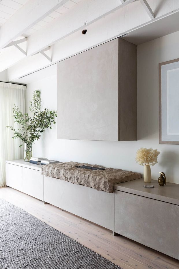 A bright, minimalist living room featuring a long low built-in media console in a warm grey textured finish, a large matching wall-mounted cabinet, white walls, pale timber flooring, and exposed white ceiling beams. A neutral textured rug, linen curtains, sculptural dried florals, and restrained decorative objects create a calm, refined interior.