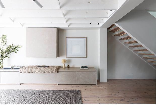 Bright, minimalist living area with white painted exposed ceiling beams, a low built-in timber-toned storage unit, neutral wall cabinetry, framed artwork, and a textured rug over wide timber floorboards. An open staircase with slim timber treads and a white structural stringer leads upward at right.