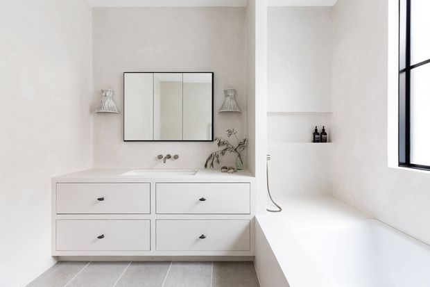 Minimalist white bathroom featuring a wide floating vanity with four drawers, integrated basin, wall-mounted mirrored cabinet, decorative wall sconces, and a built-in white bathtub beside a black-framed window. Pale textured walls and grey stone-look floor tiles create a clean, serene interior.