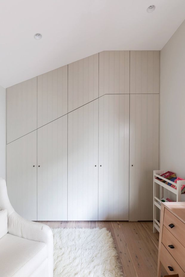 Bright, minimalist bedroom or children's room with a full-height built-in wardrobe spanning a sloped wall. The wardrobe has pale vertical timber-panelled doors with discreet dark handles and angular stepped joinery following the ceiling line. Light timber flooring, a white shag rug, a white upholstered sofa or bed edge, and a small light-coloured storage unit with books and toys are visible. Recessed ceiling lights and a soft neutral palette create an airy, calm interior.