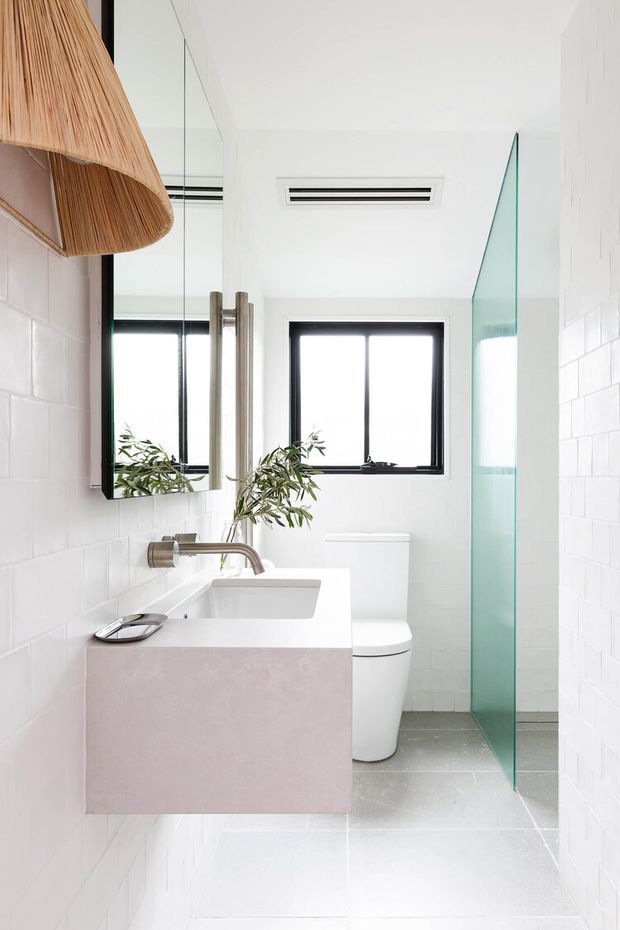 Bright, compact contemporary bathroom featuring white tiled walls and floor, a floating pale pink vanity with integrated white basin, brushed metal tapware, wall mirror, white toilet, black-framed window, and a clear green-tinted glass shower screen. A woven pendant light and leafy branch add warm natural accents.