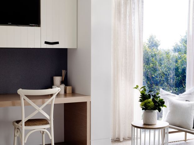 Bright, softly styled interior featuring a compact built-in home office workspace beside a large window. The space includes a pale timber desk, white built-in cabinetry, dark charcoal feature backing, a white cross-back chair, sheer curtains, a small round side table, and a white upholstered armchair with indoor plants. Natural greenery is visible outside.