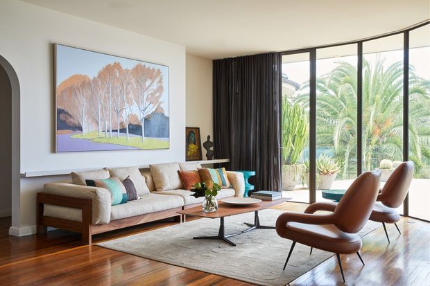 A refined living room with a low timber-framed sofa, leather lounge chairs, a slim coffee table, and a large neutral rug. Full-height black-framed glazing opens to lush palms, while a large framed artwork, dark curtains, and polished timber flooring complete the warm, elegant interior.