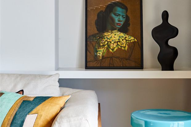 A close-up of a refined living room vignette featuring a light upholstered sofa with colourful patterned cushions, a white built-in display shelf, a framed portrait artwork, a black abstract sculptural ornament, and a glossy turquoise side table. The palette combines soft neutrals with saturated teal, mustard, and black accents.