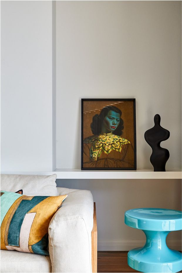 A styled living room corner featuring a white upholstered sofa with a colourful geometric cushion, a built-in recessed wall shelf displaying framed artwork and a black sculptural object, and a glossy turquoise side table. The room has warm taupe walls, white trim and shelving, and dark timber flooring.
