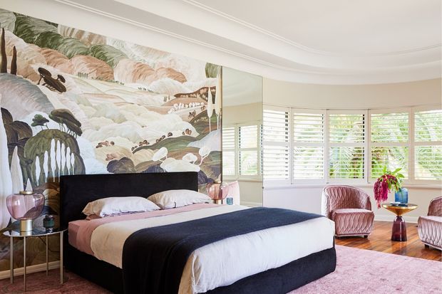 A bright, refined bedroom with a large upholstered bed dressed in layered white, blush, and dark charcoal bedding. A dramatic botanical landscape mural forms the feature wall, while plantation shutters span the curved bay windows. Two blush-toned armchairs, a small side table, decorative lighting, and a soft pink rug complete the room. The space combines classic architectural detailing with bold, luxurious decorative styling.