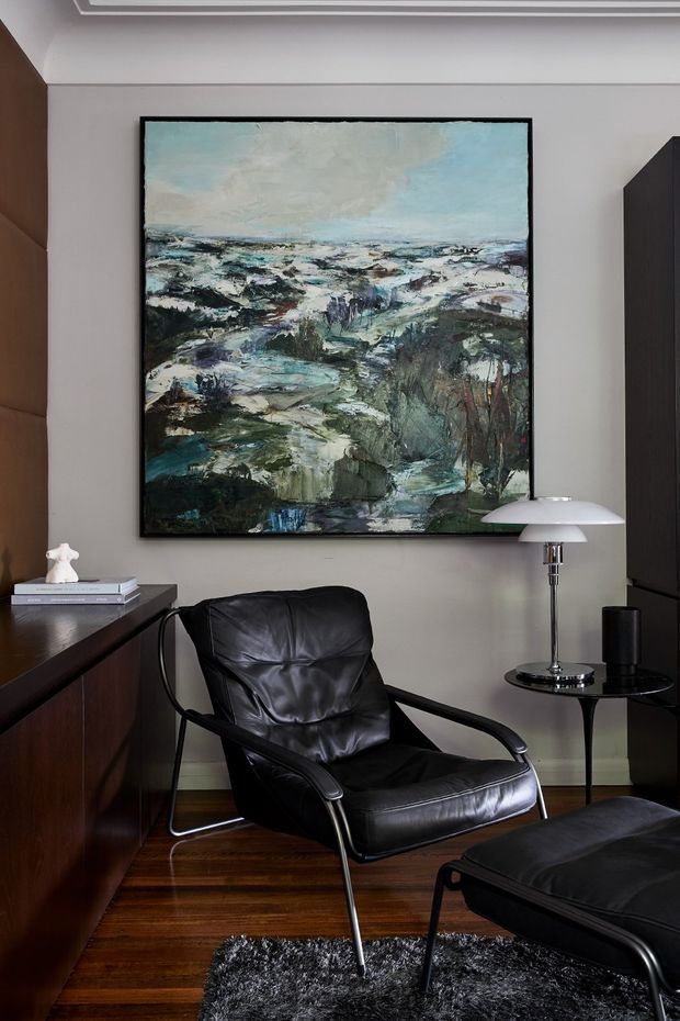 A sophisticated sitting area with a black leather lounge chair and matching ottoman, positioned beside a dark timber built-in cabinet and a small round side table with a sculptural table lamp. A large abstract landscape painting forms the focal point on the warm grey wall, with dark timber flooring, a black shag rug, and restrained mid-century-inspired furnishings. Decorative ceiling cornice detailing is visible above.