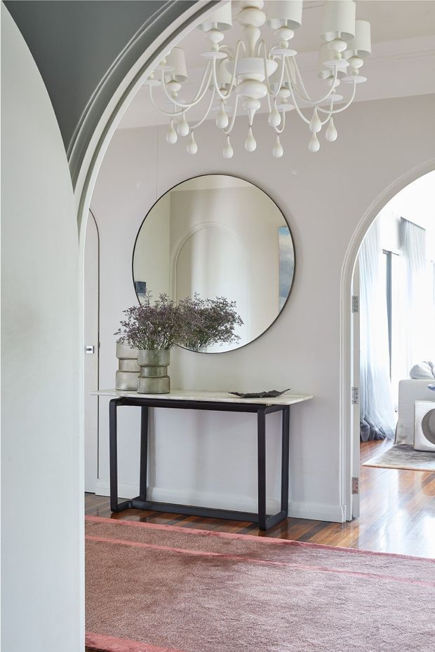 A bright, refined entry hall with curved architectural archways, white painted walls and trim, a large round wall mirror, a slim dark console table, decorative vessels, and an ornate white chandelier. A blush-toned rug covers the timber floor, while an adjoining bright living area is visible through the arched opening.