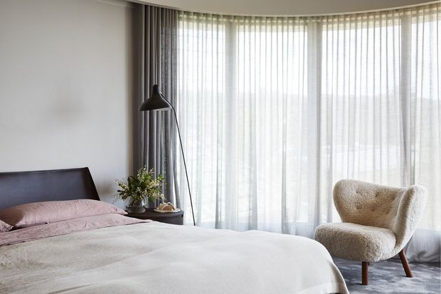 A softly lit contemporary bedroom with a low dark upholstered bed, layered neutral bedding, a tall black floor lamp, a small bedside table with greenery, and a sculptural boucle armchair. Floor-to-ceiling curved glazing is dressed with sheer white curtains and grey drapes, creating an airy, calm atmosphere.