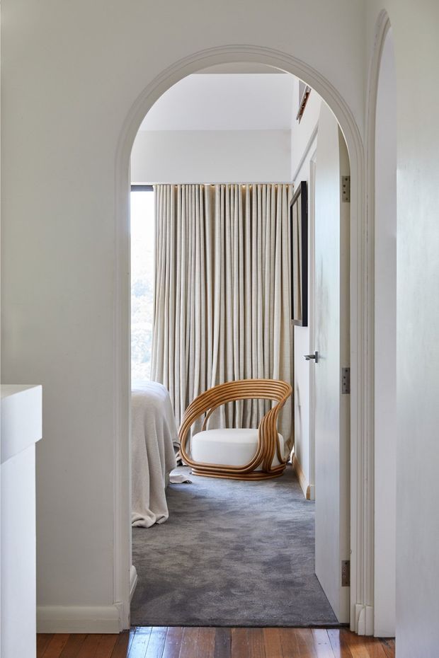View through a white-painted arched doorway into a softly furnished bedroom. The room has grey carpet, floor-to-ceiling pleated curtains, a partially visible bed, and a curved woven timber lounge chair with a white cushion. Timber flooring is visible in the foreground hallway, with simple white walls and trim throughout.