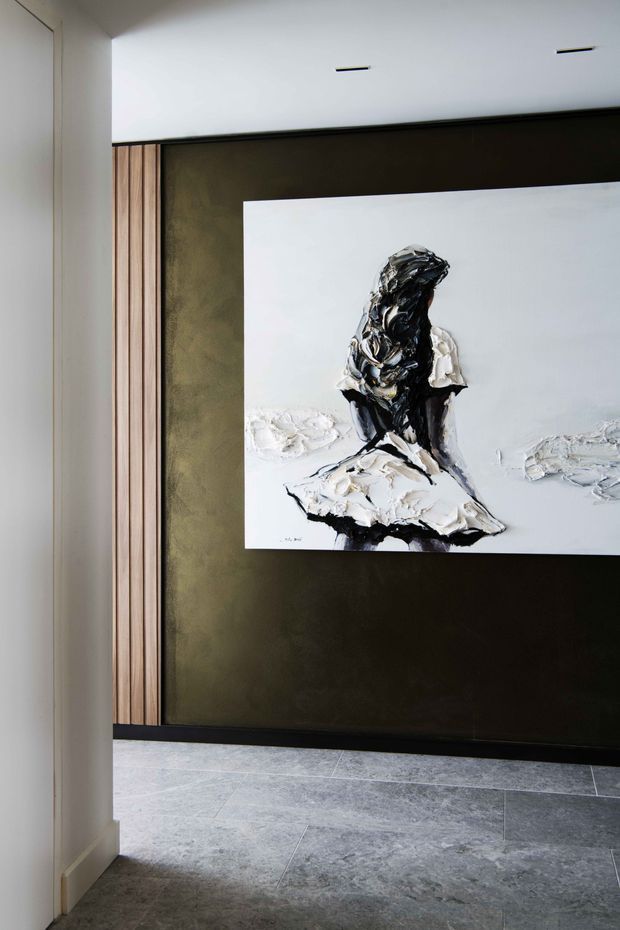 A refined interior hallway or entry area viewed through a doorway, featuring a dark textured feature wall with a large abstract monochrome artwork, vertical timber wall panelling, crisp white walls and ceiling, recessed linear ceiling lights, and large-format grey stone-look floor tiles.