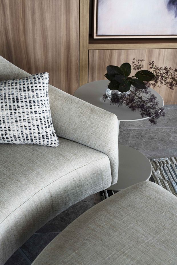 Close-up of a refined contemporary living room featuring a pale textured upholstered sofa and matching ottoman, round grey side tables, a dark stone-tiled floor, and warm timber built-in wall panelling. A small vase with foliage and framed artwork add understated decorative detail.