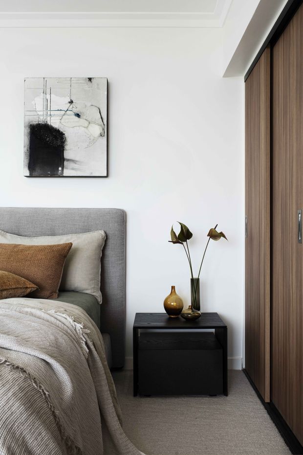 A refined bedroom vignette featuring a grey upholstered bedhead, layered neutral bedding and cushions, a black bedside table with decorative vessels, and a full-height dark timber wardrobe with black framing. White walls and detailed recessed ceiling moulding create a clean, elegant backdrop, complemented by monochrome abstract artwork and soft natural textures.