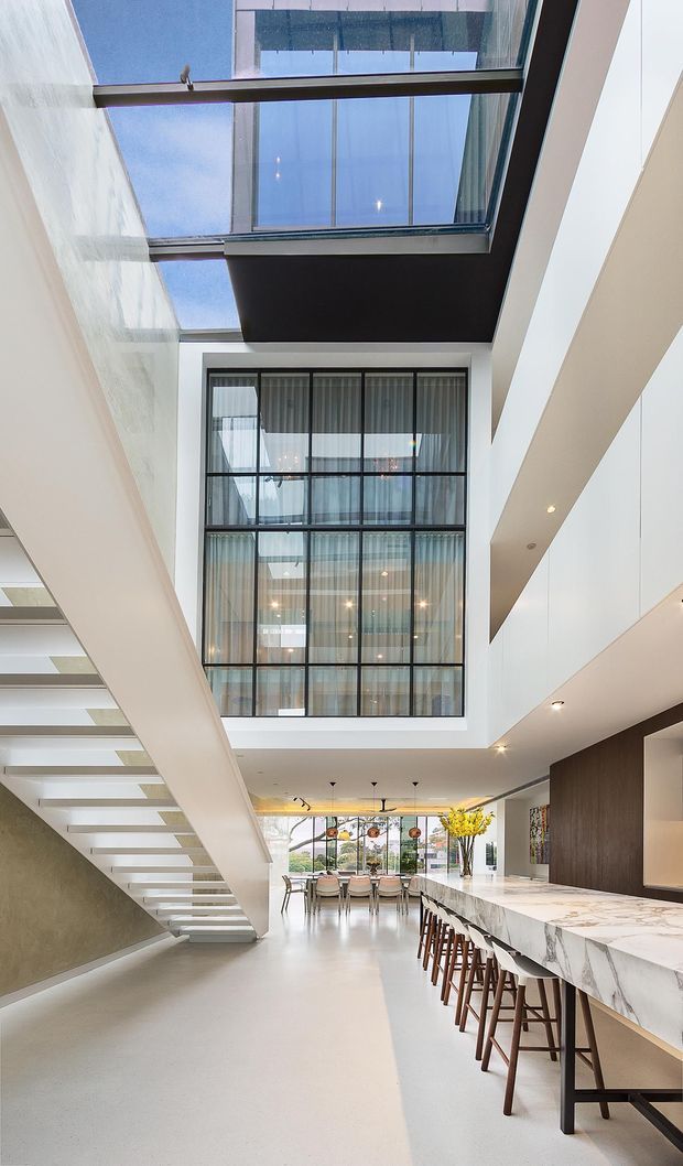 Double-height contemporary interior centered on a refined open-plan kitchen and dining area. A long white marble island with dark timber stools occupies the right side, while a sculptural open-riser staircase rises along the left. Floor-to-ceiling black-framed glazing, a large skylight, white walls, dark timber cabinetry and abundant natural light create a bright, spacious, high-end architectural interior.