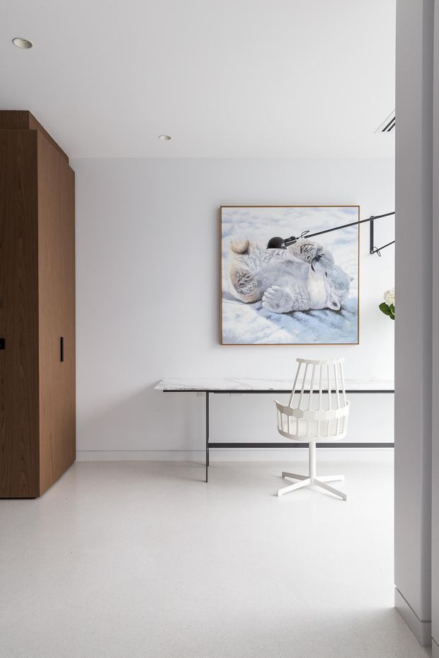 A bright, minimalist interior view showing a compact entry or hallway area. A tall dark timber built-in wardrobe or storage unit stands on the left, with a slim marble-topped console table against a white wall. A white sculptural chair, black articulated wall lamp, framed artwork, recessed ceiling lights, and pale flooring complete the restrained monochromatic space.