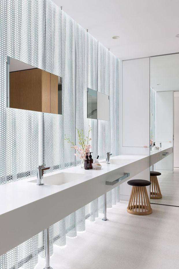 A bright contemporary bathroom with a long floating white double vanity, integrated basins, chrome wall-mounted taps, large mirrors, and timber stools. A perforated metal screen spans the full-height window wall, creating a distinctive patterned backdrop with abundant diffused daylight. The space has pale smooth flooring, white cabinetry, and minimalist detailing.