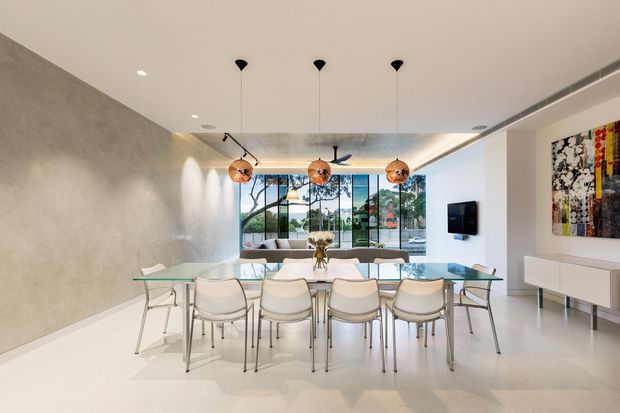 Bright contemporary open-plan dining area with a long glass dining table and multiple white upholstered chairs, opening toward a living space and large floor-to-ceiling windows. The room features a smooth concrete-look feature wall, pale flooring, recessed lighting, three copper pendant lights, a ceiling fan, minimalist white cabinetry, and colourful wall art.
