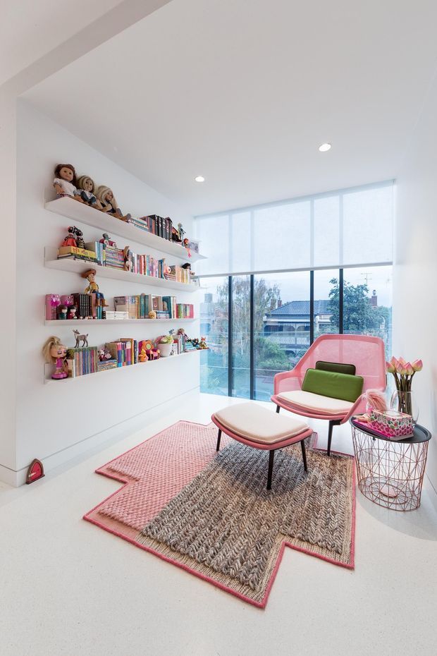 Bright, playful reading nook or children’s room with wall-mounted open shelving filled with books, dolls, and toys. A pink upholstered lounge chair and matching ottoman sit beside a small wire-frame side table, with layered pink and textured neutral rugs. Floor-to-ceiling windows with roller blinds provide abundant natural light and views of neighbouring homes.