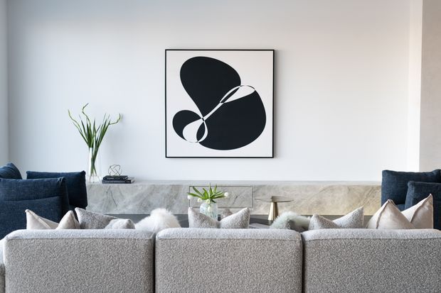 A refined contemporary living room viewed from behind a large light-grey sectional sofa. The room features a pale stone or marble fireplace surround and mantel, deep navy armchairs at either side, neutral cushions, a minimalist black-and-white abstract artwork, and a simple glass vase with greenery. The palette is calm and predominantly white, grey, navy, and black.