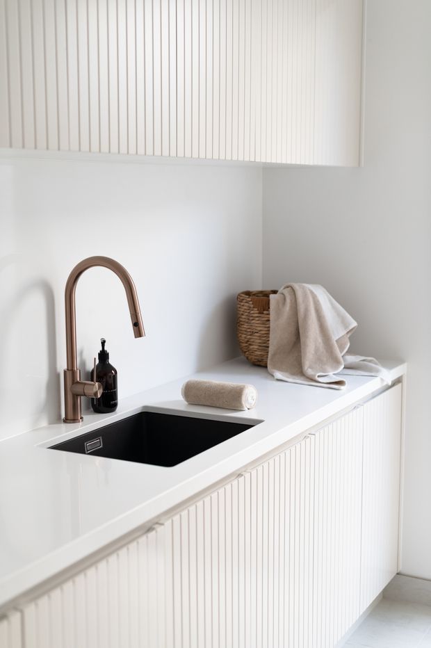 Bright, minimalist laundry or utility room with white vertical fluted cabinetry, a white benchtop, and a deep black undermount sink. A curved brushed bronze tap, dark soap dispenser, woven basket, and folded neutral towels add subtle warmth and contrast. Light flooring and white walls create a clean, calm appearance.