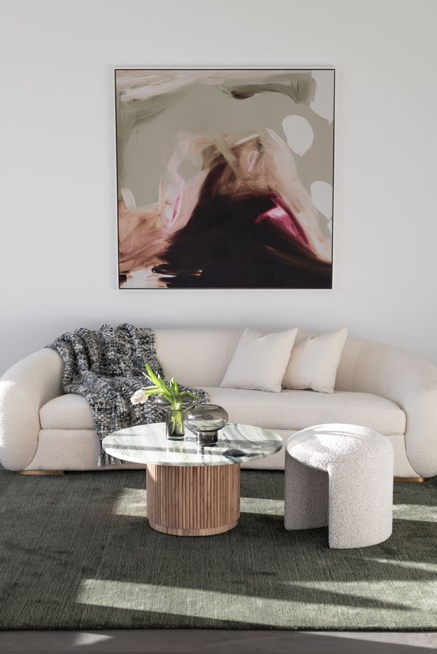 A softly lit contemporary living room featuring a curved cream boucle sofa, textured dark rug, round marble-topped coffee table with a fluted timber base, and a matching rounded boucle ottoman. An abstract artwork hangs on the light wall above the sofa, with a small vase arrangement and decorative bowl on the table. Strong sunlight creates patterned shadows across the rug and furniture.
