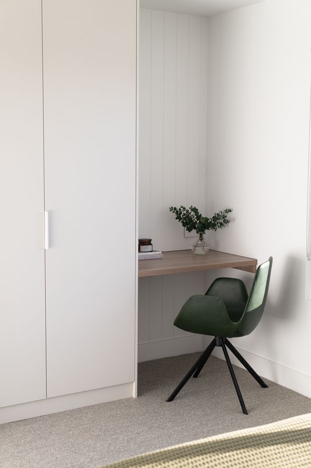 A compact, minimalist home office nook with a built-in white storage cupboard, a wall-mounted timber-look desk, vertical white wall panelling, and a dark green swivel chair. Neutral carpet, soft natural light, and a small vase of greenery create a calm, understated workspace.