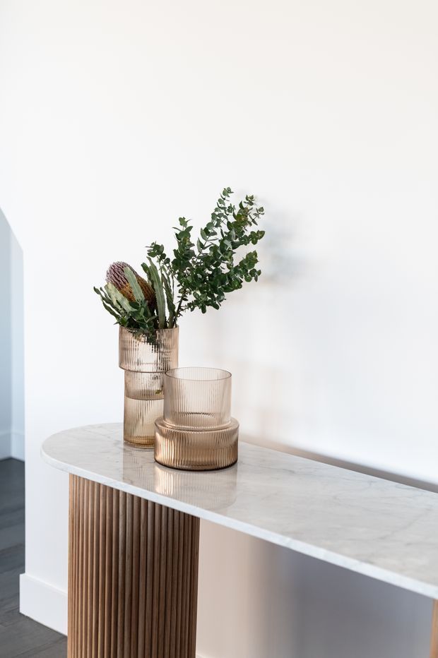 A close-up interior vignette of a slim console table against a white wall. The console has a white marble or stone-look top, a vertically fluted warm wood pedestal base, and rounded ends. Ribbed amber glass vessels and a tall vase of dried and leafy botanical stems sit on top. A dark grey floor and white skirting are visible.