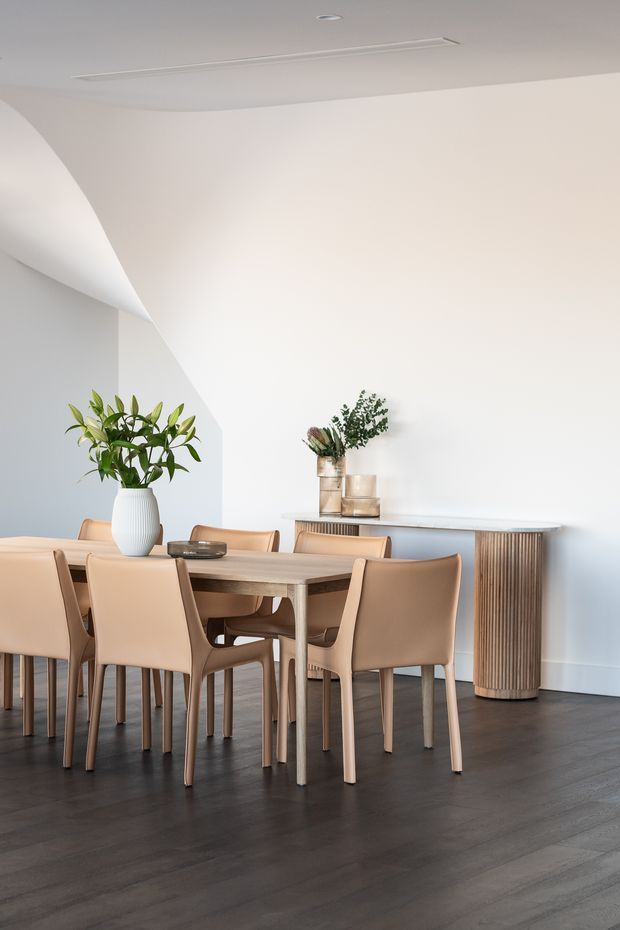 Bright, refined dining room with a light neutral palette, dark timber flooring, a contemporary rectangular dining table surrounded by upholstered tan dining chairs, and a fluted timber sideboard against a white wall. Sculptural curved ceiling detailing and recessed linear lighting add architectural interest, while simple botanical arrangements provide natural accents.