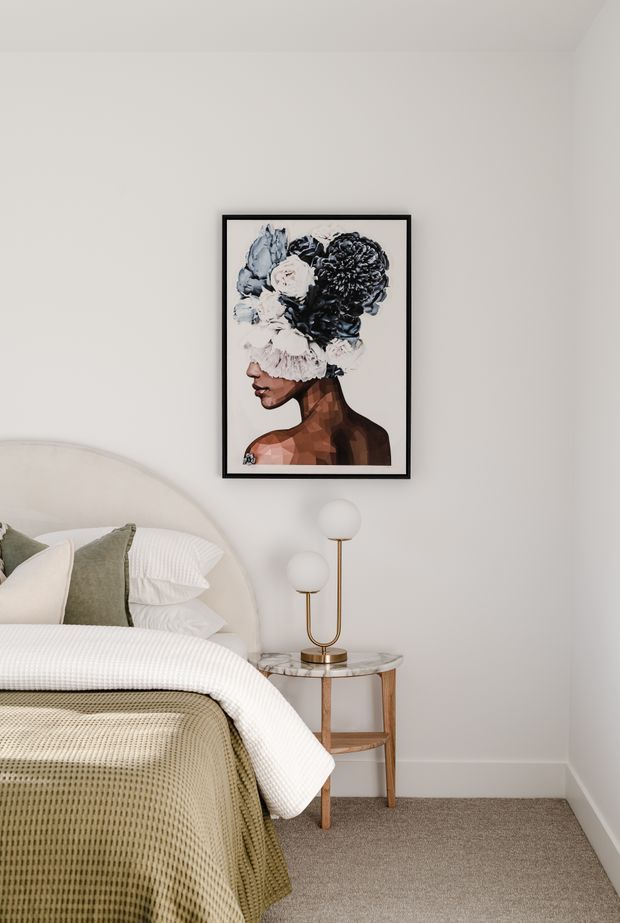 A softly styled bedroom corner with a curved upholstered bedhead, layered white and sage-green bedding, a marble-topped timber bedside table, and a sculptural brass-and-glass double globe lamp. A framed monochrome portrait artwork hangs above the table. The room has warm neutral carpeting, pale walls, and a calm, refined palette.