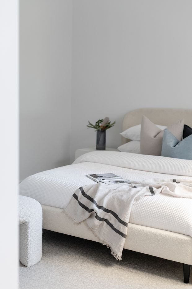 A softly lit contemporary bedroom featuring a low upholstered bed with a rounded headboard, layered white bedding, pale neutral and blue cushions, a textured throw with dark stripes, and a small dark vase on a bedside surface. The room has light grey walls, pale carpet, and a calm minimalist presentation.