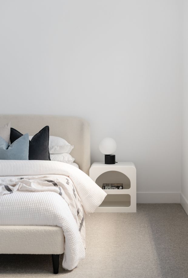 A calm, minimalist bedroom vignette featuring a softly upholstered cream bed with layered white bedding and muted grey-blue cushions. A sculptural white bedside table with rounded openings holds a small black lamp with a spherical white shade. The room has plain light walls, soft grey carpet, and restrained neutral styling.