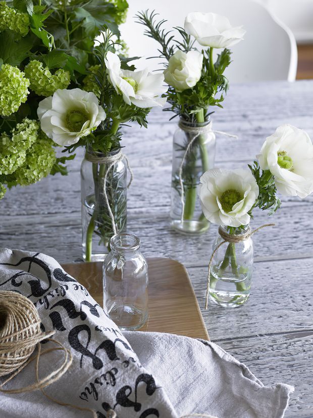A styled tabletop scene with several small glass jars holding white flowers and green foliage, arranged on a weathered grey timber table. A pale timber serving board, woven basket, patterned black-and-white textile, and white fabric create a soft rustic decorative composition. A white dining chair is partially visible in the background.
