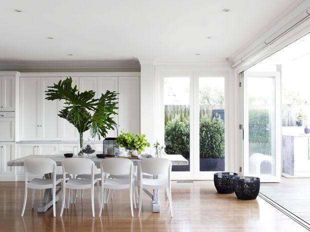 Bright open-plan dining area with a long white dining table and matching white chairs, positioned beside a white built-in kitchen with paneled cabinetry. Large glazed doors open to a planted outdoor area, while pale timber flooring, white walls, recessed ceiling lights, and indoor plants create an airy, light-filled space.
