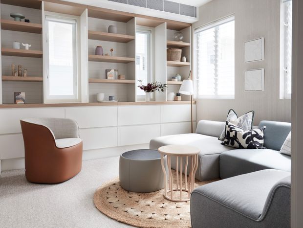 Bright, refined living room with built-in white cabinetry and open timber shelves spanning the back wall, integrated around tall windows. A pale grey sectional sofa, sculptural brown armchair, round nesting tables, floor lamp, and textured circular rug create a calm contemporary interior. Soft daylight enters through large windows with white shutters or blinds.