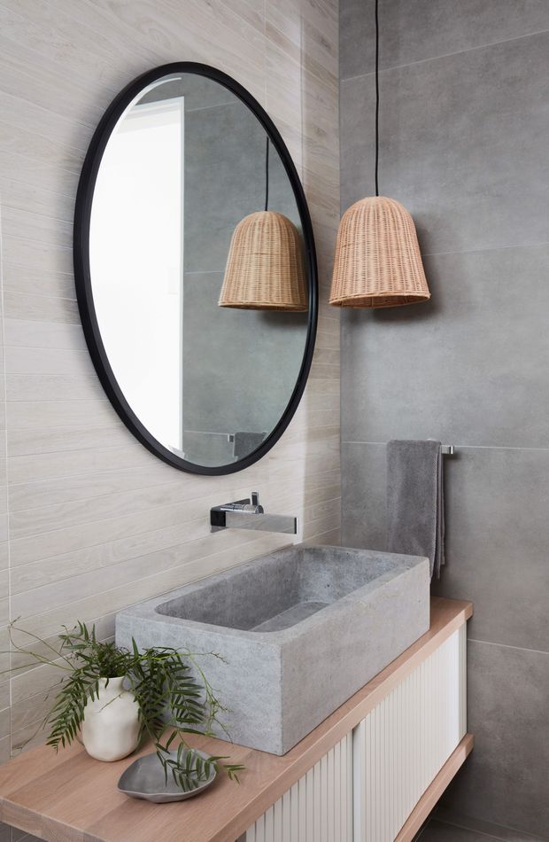 A refined contemporary bathroom featuring a freestanding rectangular concrete vessel basin on a timber vanity with white fluted cabinetry. Large grey concrete-look wall tiles contrast with pale wood-look tiles, while a black-framed oval mirror and woven pendant light add warmth and texture. A chrome wall-mounted tap, grey towel and small potted plant complete the setting.