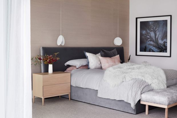 A softly styled contemporary bedroom with a low upholstered bed, dark padded headboard, pale timber feature wall, matching pendant lights, light bedding, a fuzzy throw, bedside table, framed artwork and a small upholstered bench. The room has a calm, muted neutral palette and abundant filtered daylight.