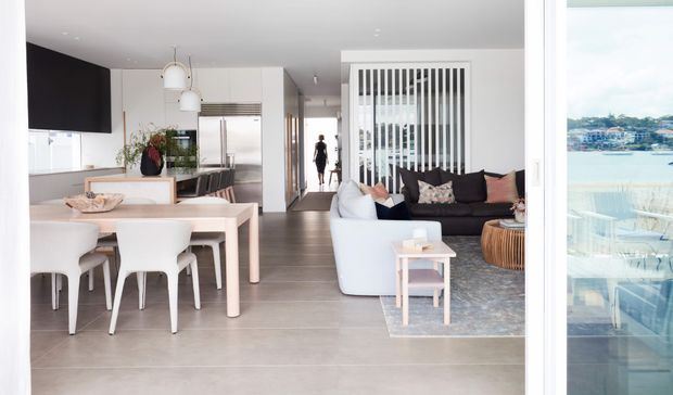 Bright open-plan contemporary apartment interior with a dining area and kitchen on the left and a living room on the right. The space features pale grey tiled flooring, white cabinetry and furniture, dark feature cabinetry or wall panel, a black sofa, pendant lights, and a vertical white slatted room divider. Large glazing at right provides a water view.