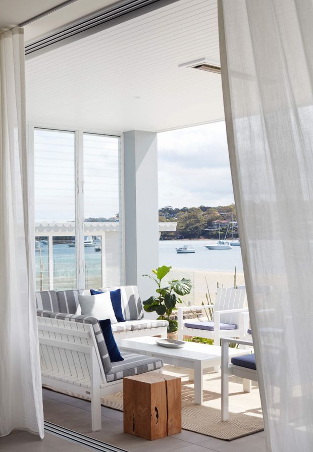 Bright coastal-style enclosed outdoor lounge or sunroom opening toward a sandy waterfront, furnished with white-painted timber seating, striped blue-and-white cushions, a white coffee table, and sheer curtains. Large glazed openings provide expansive views across the water, with a relaxed, airy atmosphere and abundant natural light.