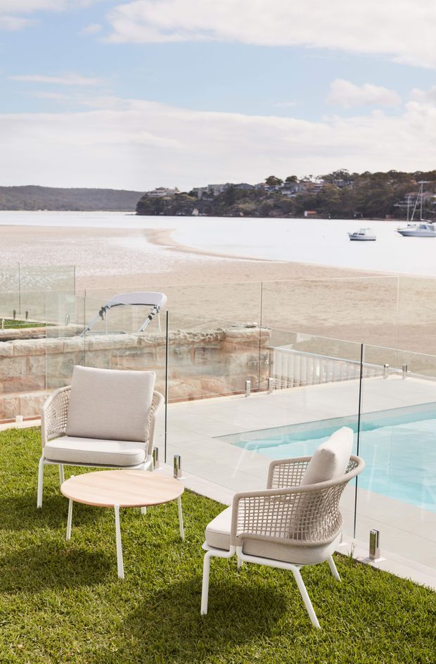 A bright outdoor terrace overlooking a sandy beach and calm bay, with a swimming pool, frameless glass balustrade, two upholstered woven lounge chairs, and a small round table on a manicured lawn. Boats and a wooded residential shoreline are visible in the distance.